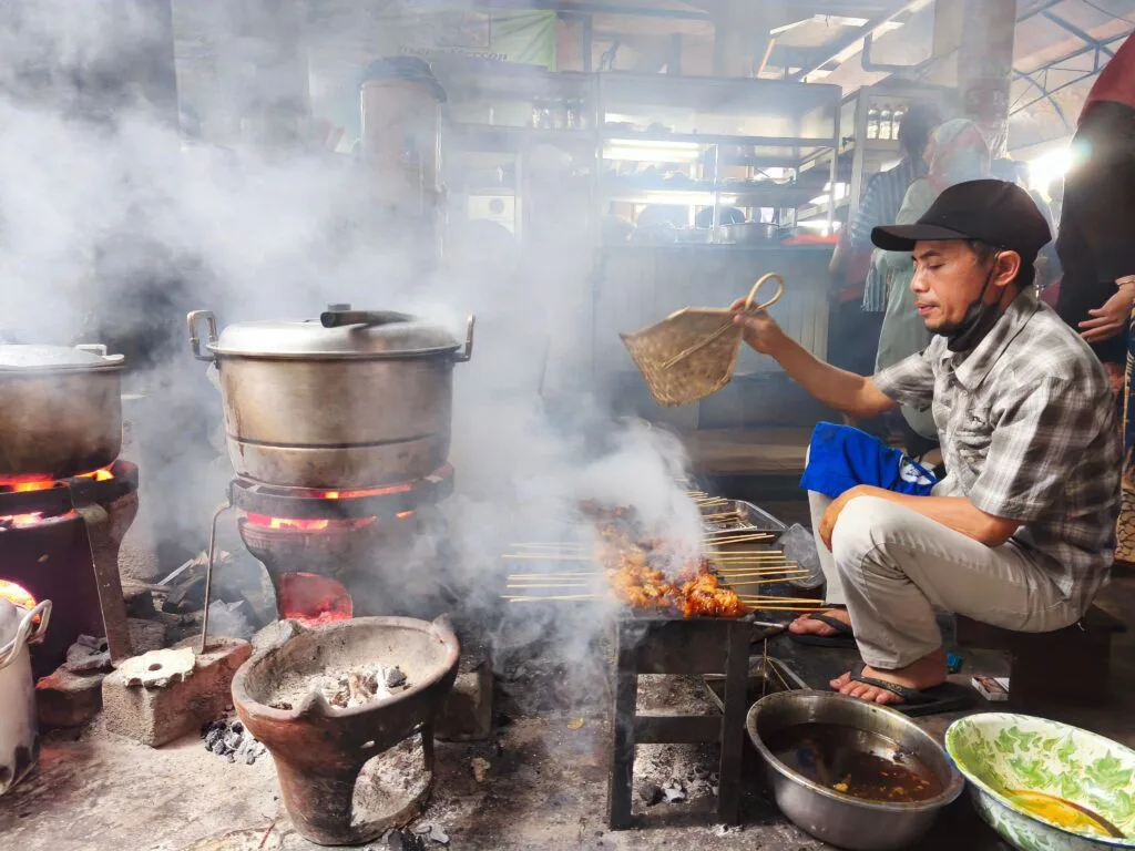 Street food vendor grills satay on skewers over open flames in a traditional market in Yogyakarta.
